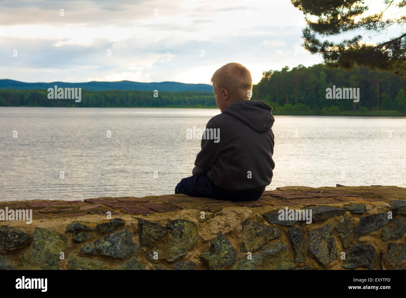 trauriges Kind sitzt allein am See an einem bewölkten Tag Stockfoto