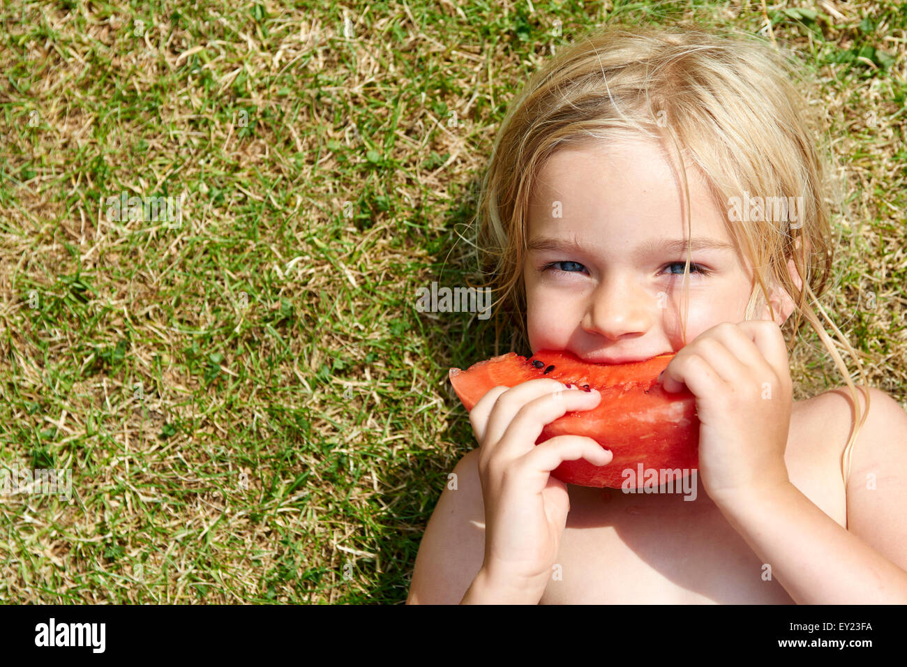 Porträt von kleinen Kind blondes Mädchen Essen Stück Wassermelone, liegend auf grünem Rasen, Sommer, Entspannung Stockfoto