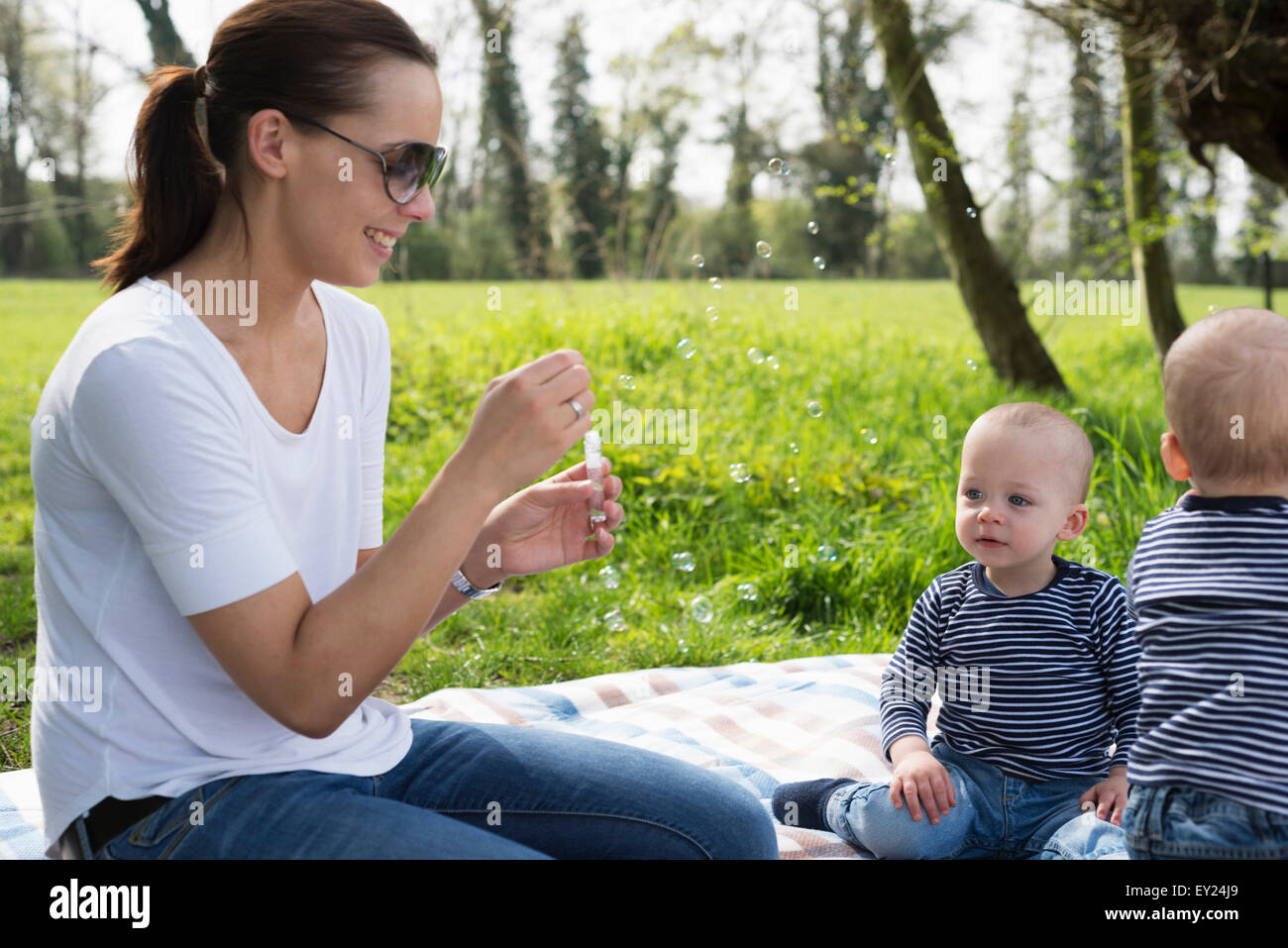 Junge Mutter bläst Seifenblasen für Baby-Zwillingen auf der Picknickdecke Stockfoto