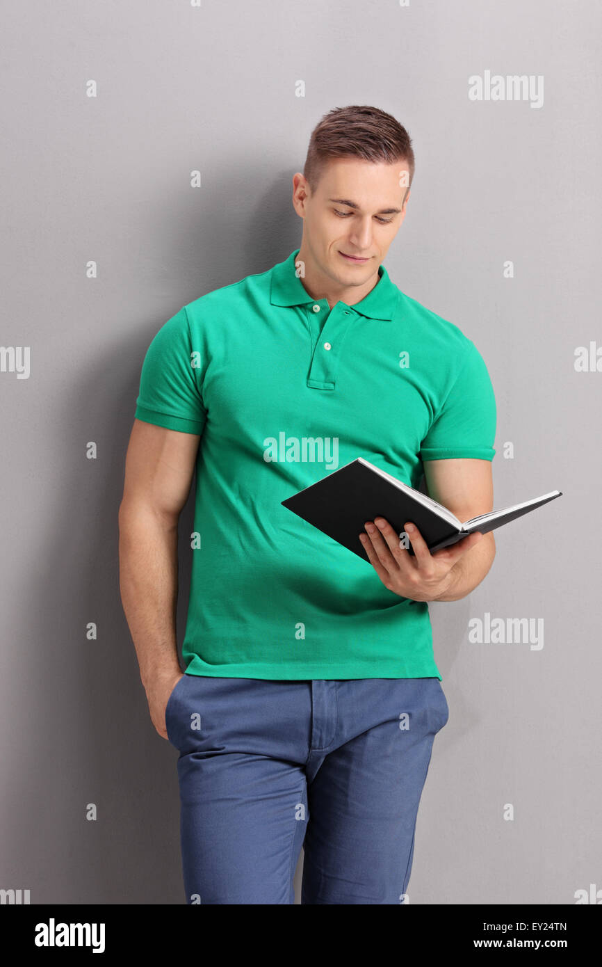 Vertikale Aufnahme eines Jünglings lässig in ein grünes Hemd, ein Buch lesen und stützte sich auf eine graue Wand Stockfoto