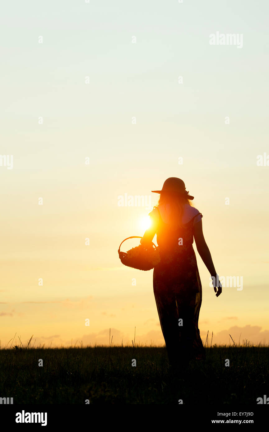 Teenager-Mädchen tragen ein Sommer Hut zu Fuß mit einem Korb voller Blumen bei Sonnenuntergang. Silhouette Stockfoto