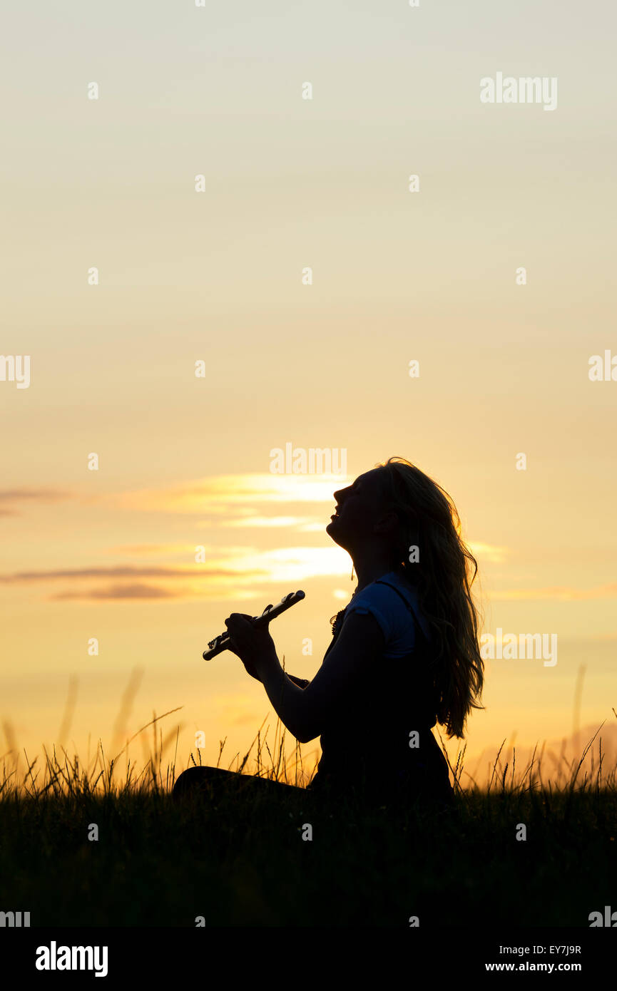 Teenager-Mädchen saß auf dem Rasen ein Flötenspiel bei Sonnenuntergang. Silhouette Stockfoto