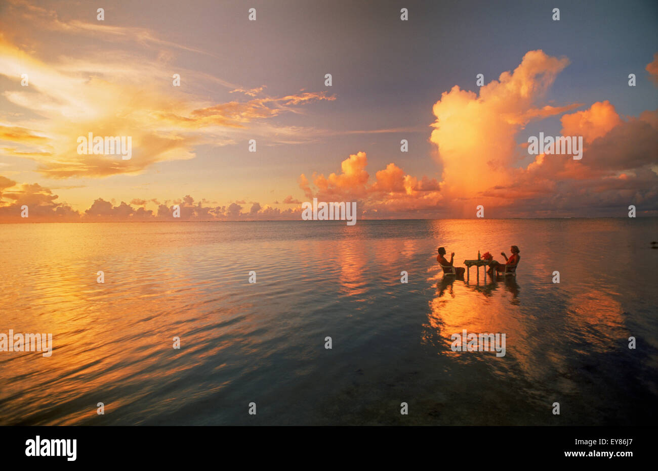 Pärchen, die am Tisch sitzen und einen Drink bei Sonnenuntergang in der Lagune inmitten des Pazifischen Ozeans genießen. Cook-Inseln. Urlaub Ruhe und Einsamkeit im Paradies Stockfoto