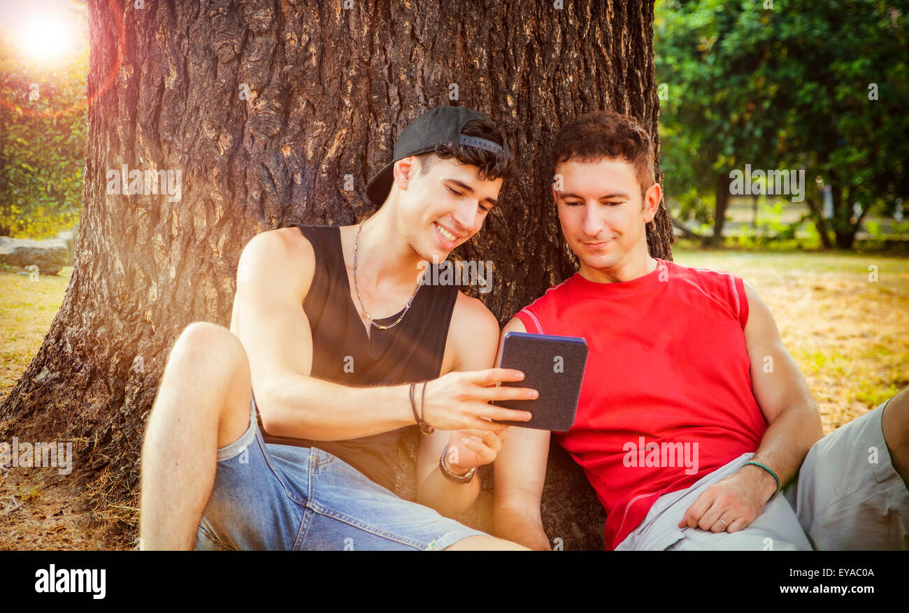 Zwei junge männliche Freunde mit lässig trendige Sommerkleidung stützte sich auf einen alten Baumstamm, während gerade etwas lustiges auf einem Tisch Stockfoto