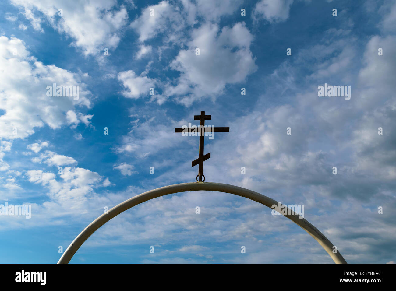 Weißen halbkreisförmigen Bogen von Eisenrohr mit dunklen orthodoxen Kreuz darauf gegen den blauen Himmel mit Wolken. Stockfoto