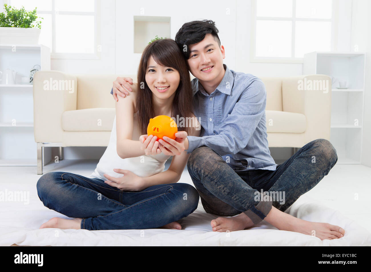 Junger Mann und schwangere Frau sitzen auf dem Boden und halten Piggy Bank zusammen, Stockfoto