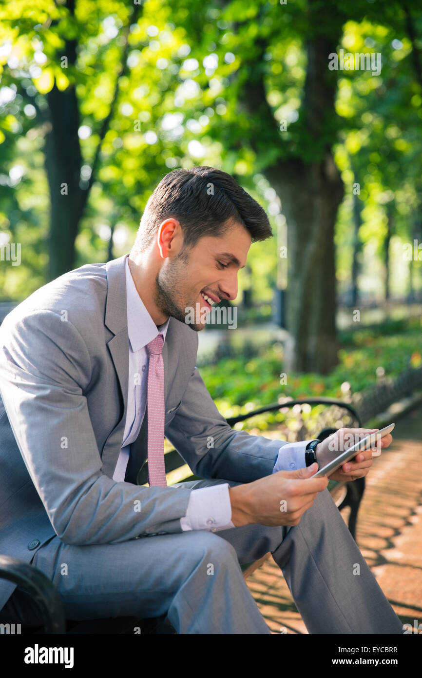 Lächelnde junge Usinessman sitzen auf der Bank im Freien im Stadtpark und Tablettcomputer Stockfoto