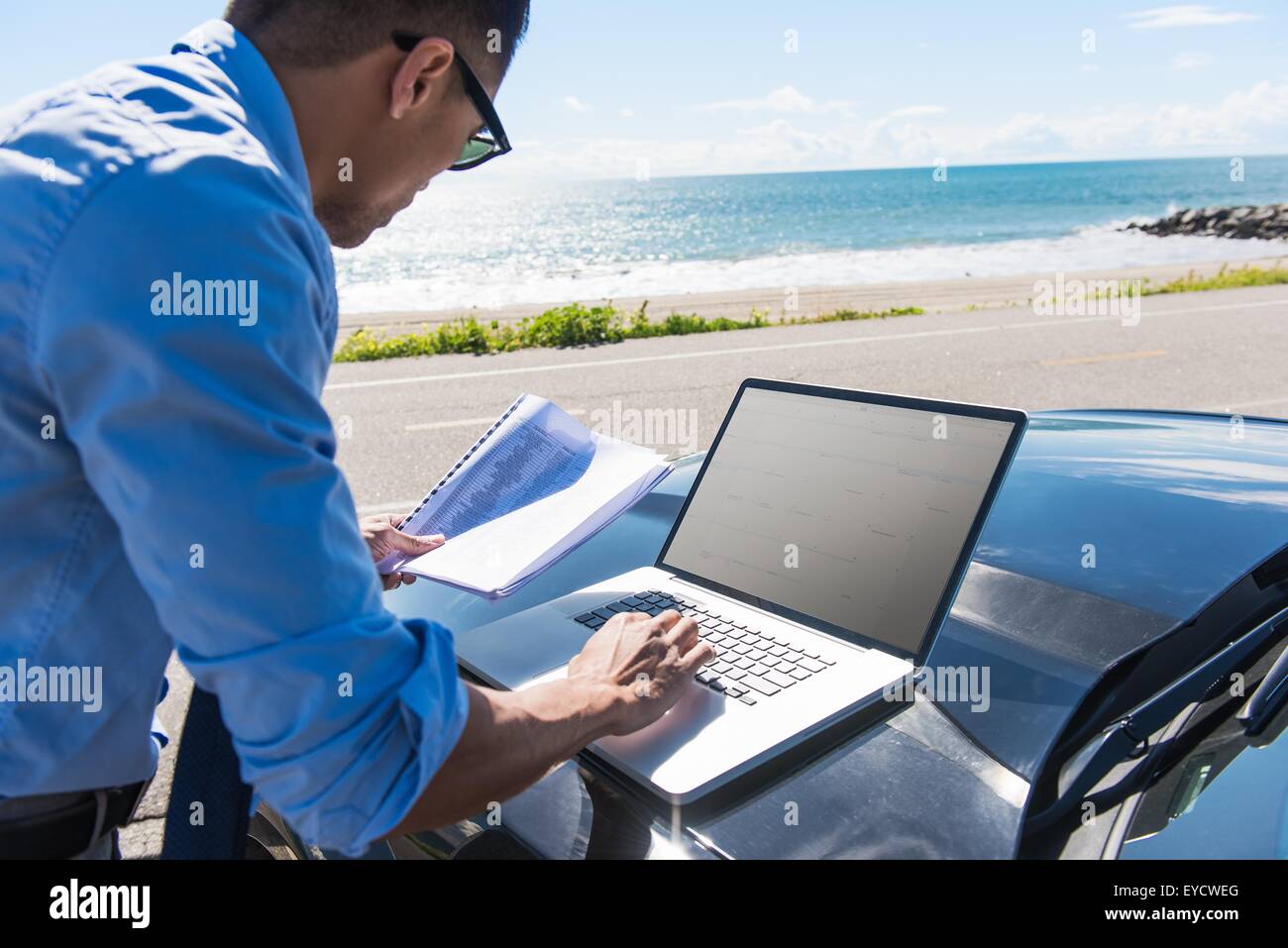 Junger Geschäftsmann auf Küste Parkplatz auf Laptop tippen, während der Lektüre Papierkram Stockfoto