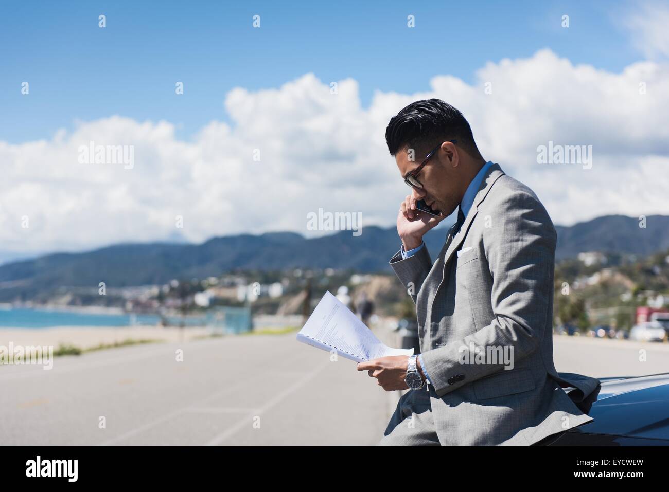 Junger Geschäftsmann sitzt auf Motorhaube auf küstennahen Parkplatz am Smartphone sprechen Stockfoto