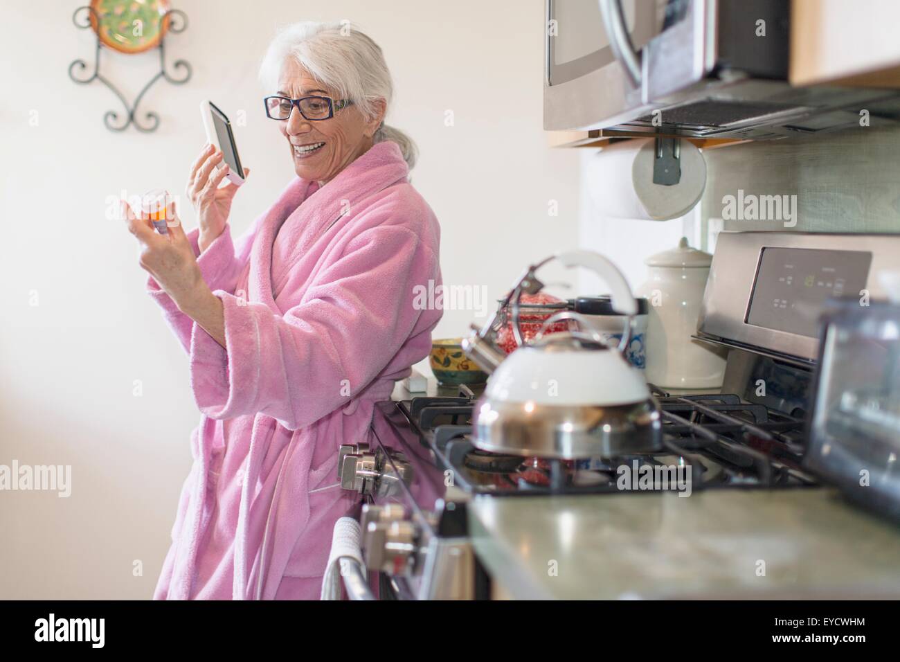 Ältere Frau im Küche Rezept halten Drogen Flasche, während auf Smartphone sprechen Stockfoto