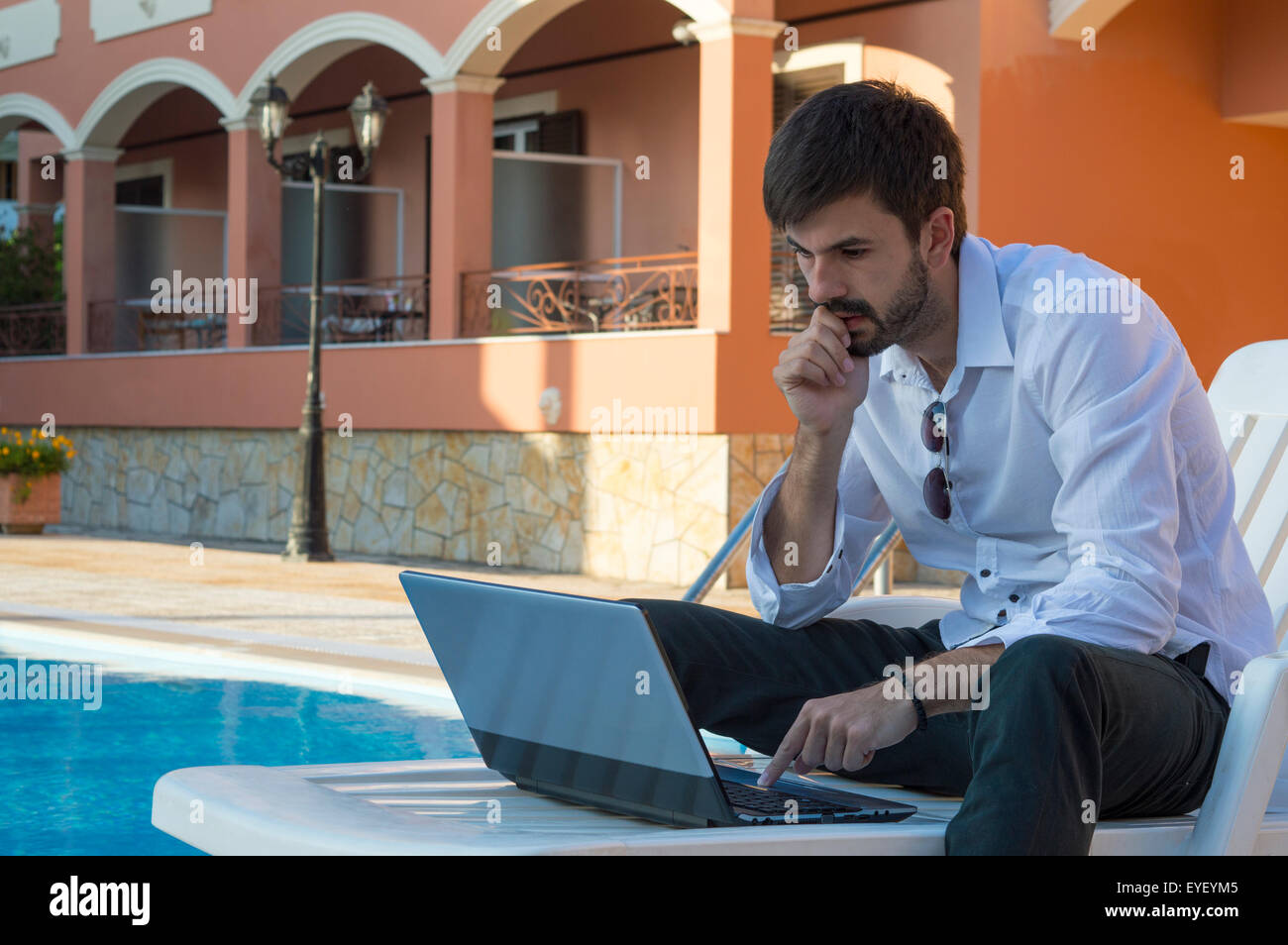 Junger Geschäftsmann arbeitet an seinem Laptop am Pool im Liegestuhl Stockfoto