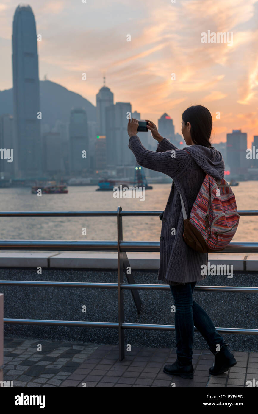 Eine junge Frau nimmt ein Bild mit ihrer Kamera den Hafen und die Skyline von Hong Kong bei Sonnenuntergang, Kowloon; Hongkong, China Stockfoto