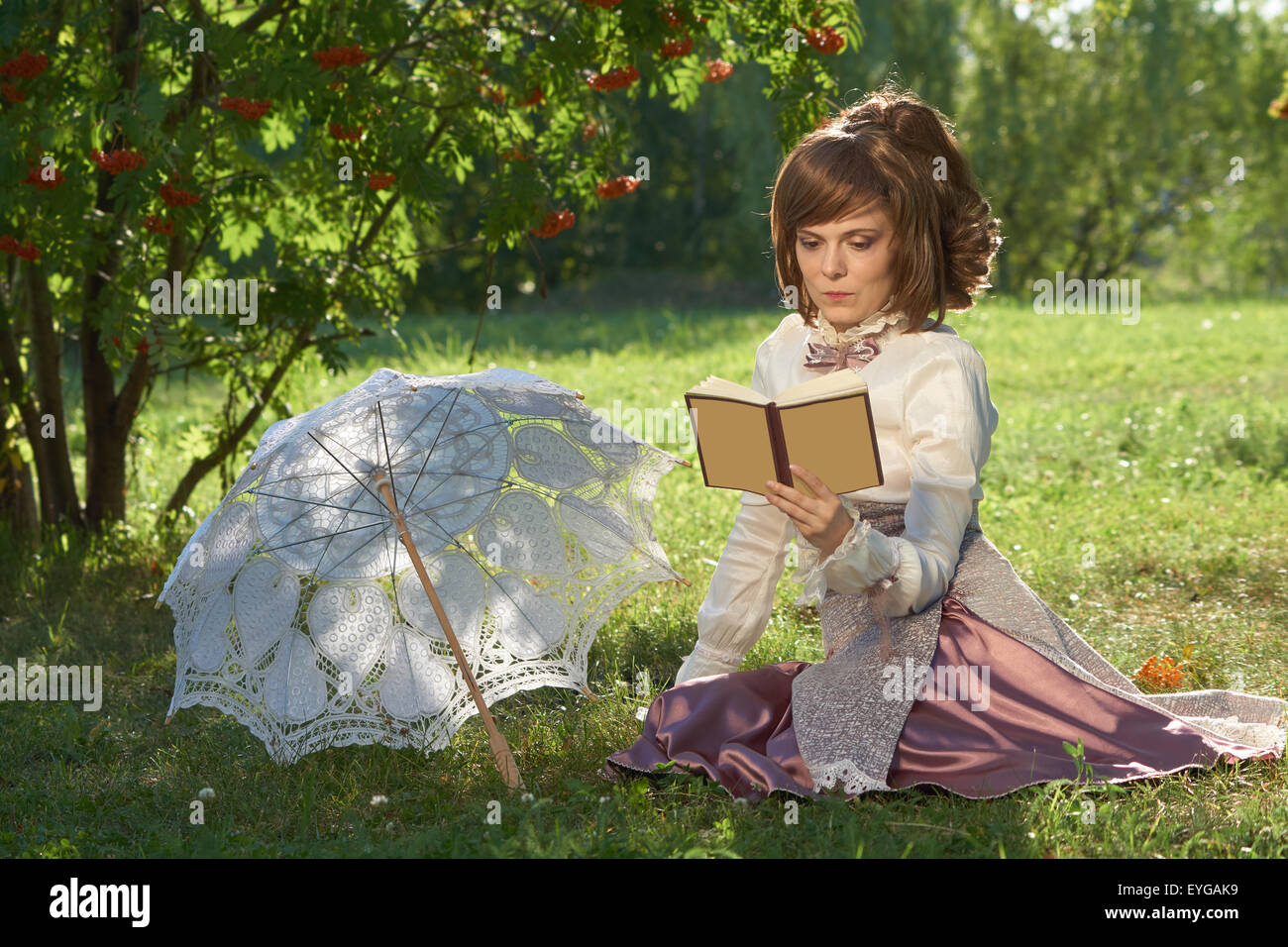 Mädchen im Retro-Stil Kleid liest Buch im Park mit Sonnenschirm neben ihr Stockfoto