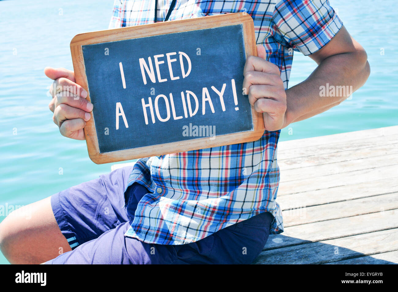 ein junger kaukasischer Mann sitzen in einem hölzernen Pier zeigt eine Tafel mit dem Text, den ich einen Urlaub in es geschrieben brauche Stockfoto