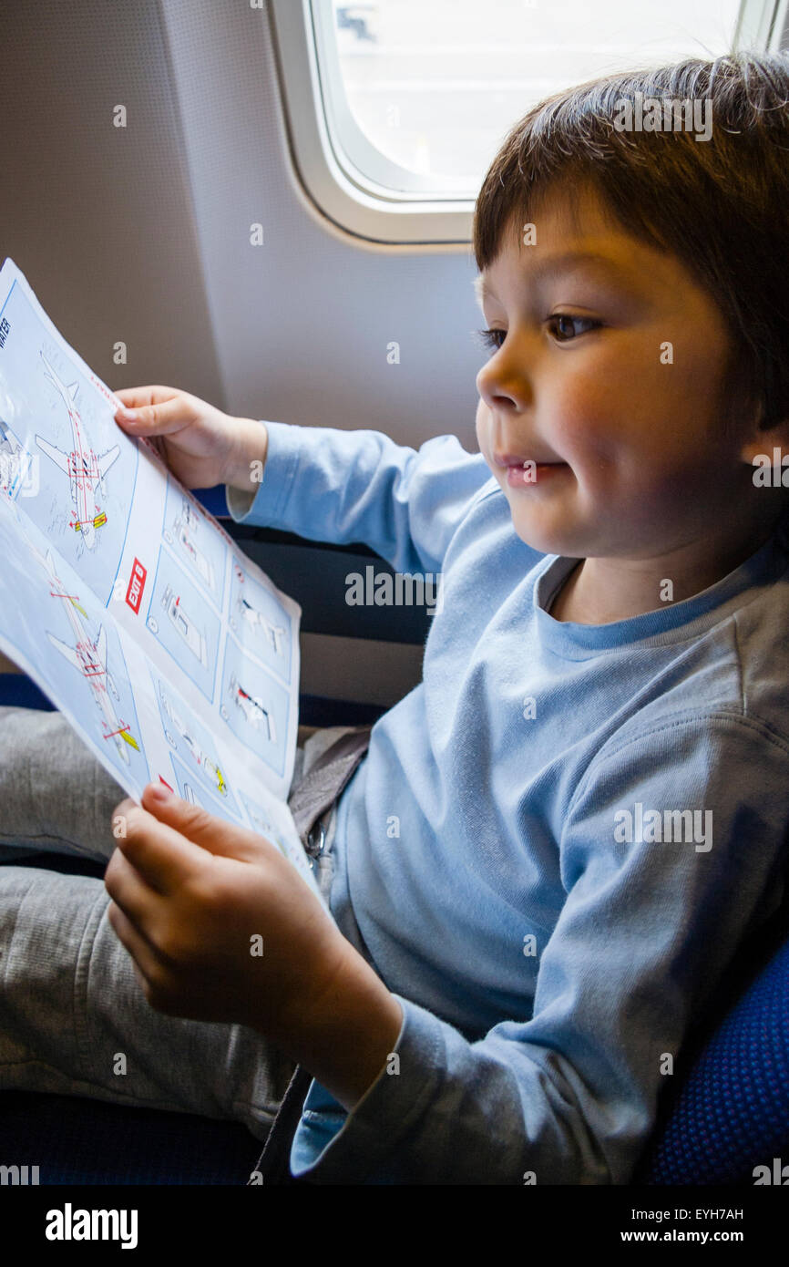 Junge sitzt im Flugzeug Fenster Beifahrersitz und lesen die Sicherheit der Gebrauchsanweisung. Kaukasier Kind, 4-5 Jahre alt. Hält Karte mit beiden Händen. Stockfoto