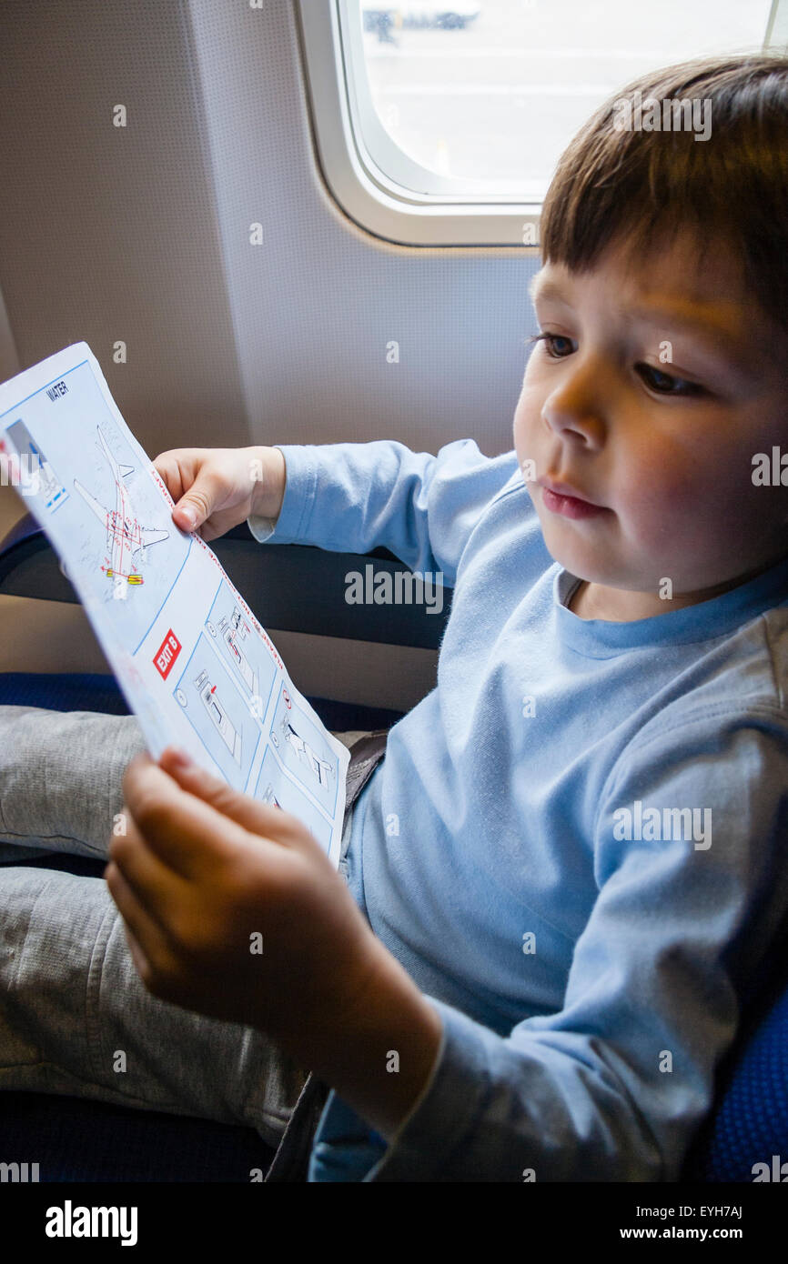 Junge sitzt im Flugzeug Fenster Beifahrersitz und lesen die Sicherheit der Gebrauchsanweisung. Kaukasier Kind, 4-5 Jahre alt. Hält Karte mit beiden Händen. Stockfoto