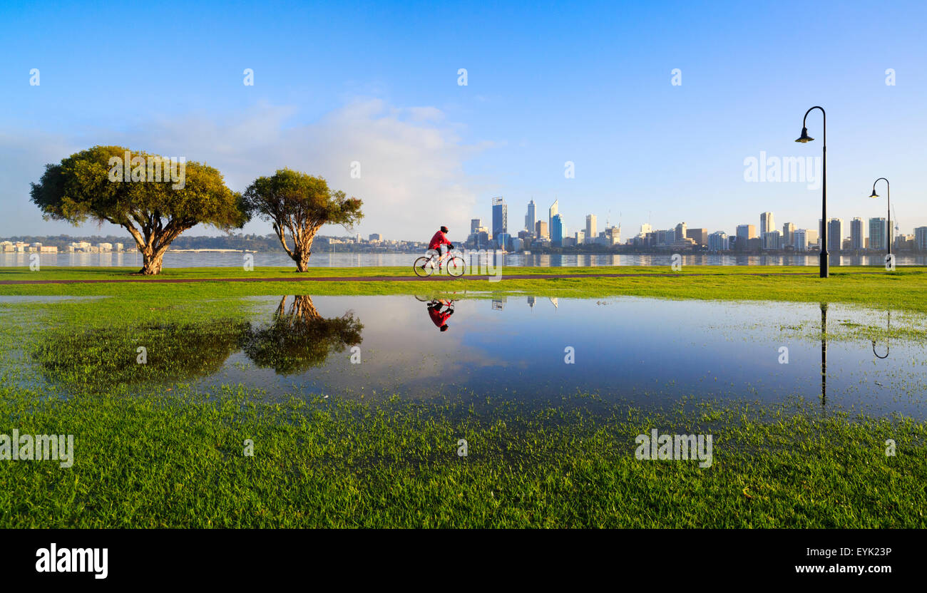 Ein Mann, ein Radweg radeln spiegelt sich in einer Pfütze mit der Stadt im Hintergrund.  Perth, Western Australia, Australia Stockfoto