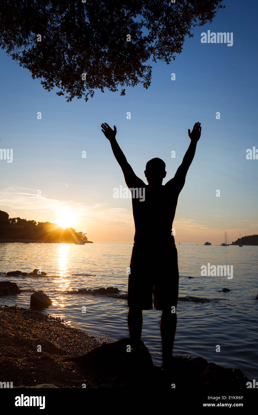 Ein Mann steht auf einem Felsen an der Küste mit Blick auf das offene Meer und grüßte die Sonne mit erhobenen Armen in Rovinj, Kroatien. Stockfoto
