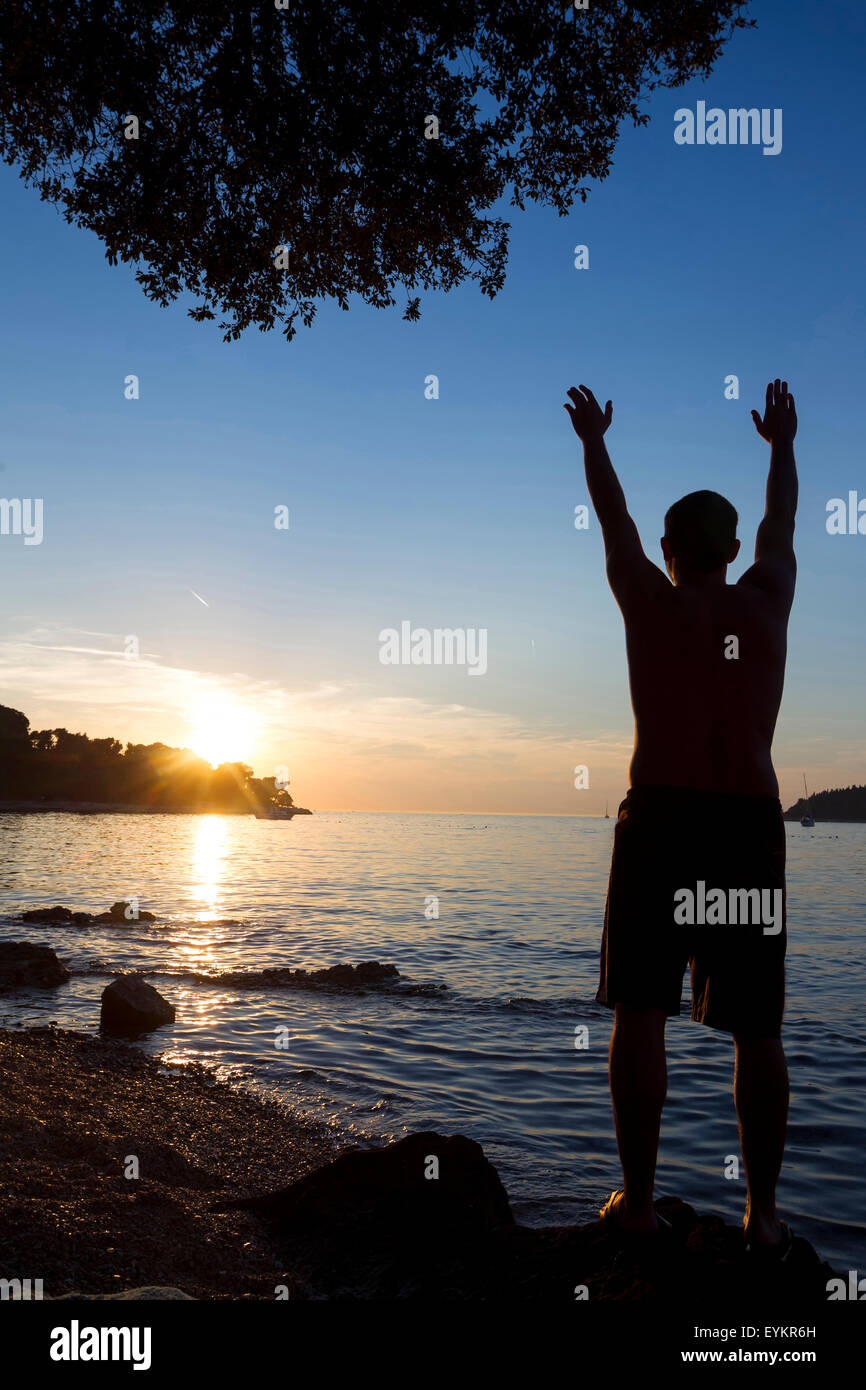 Ein Mann steht auf einem Felsen an der Küste mit Blick auf das offene Meer und grüßte die Sonne mit erhobenen Armen in Rovinj, Kroatien. Stockfoto