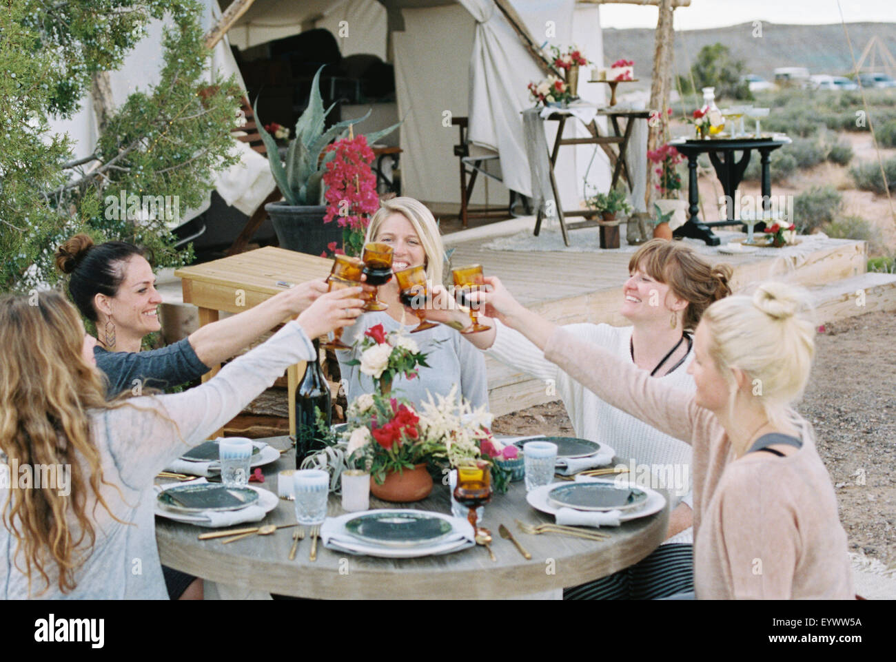Eine Gruppe von Frauen genießen eine Mahlzeit im Freien durch ein großes Zelt in einer Wüstenlandschaft, einen Toast durch klirrende Gläser. Stockfoto