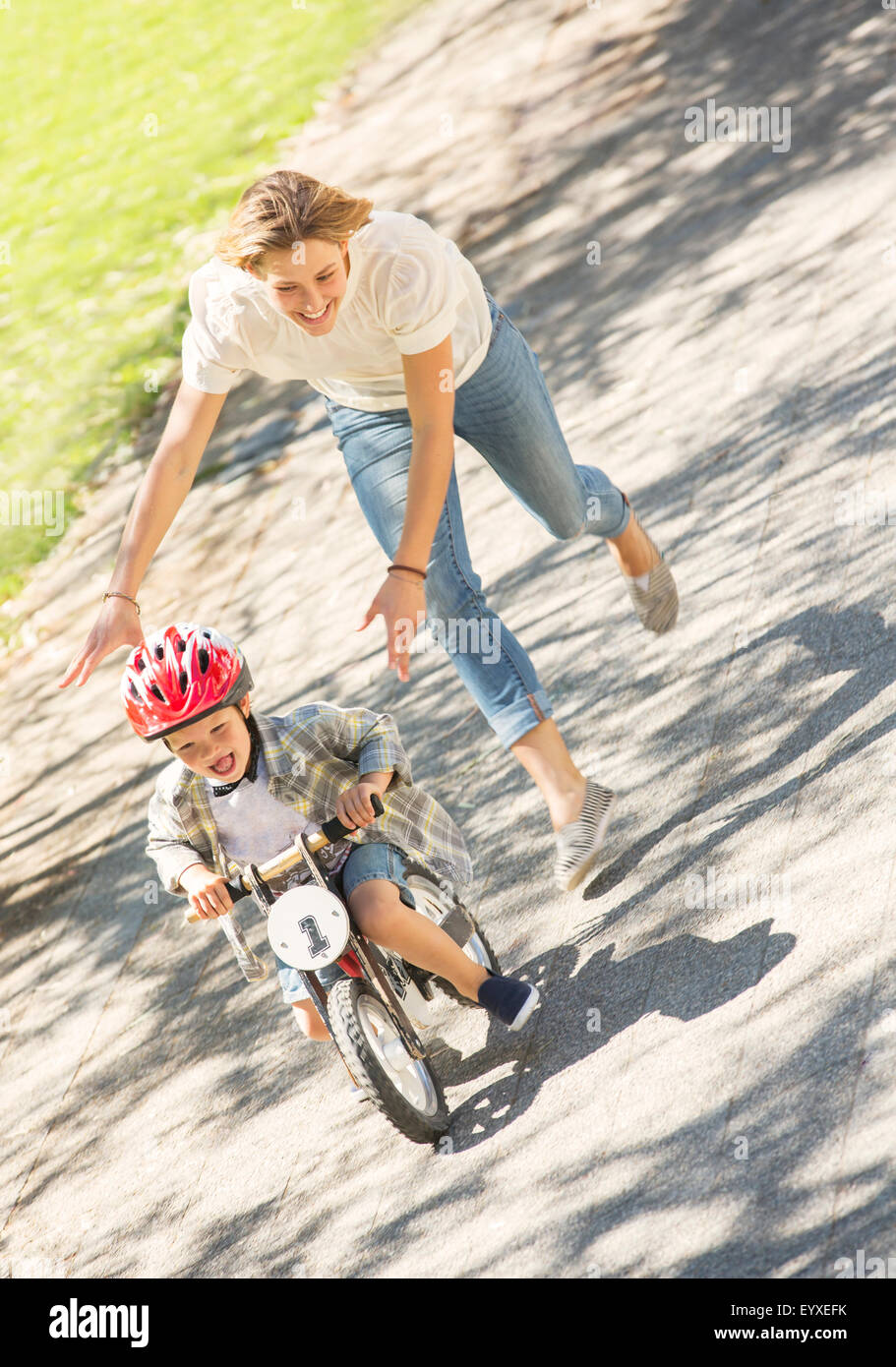 Mutter drängt Sohn mit Helm auf dem Fahrrad im sonnigen park Stockfoto