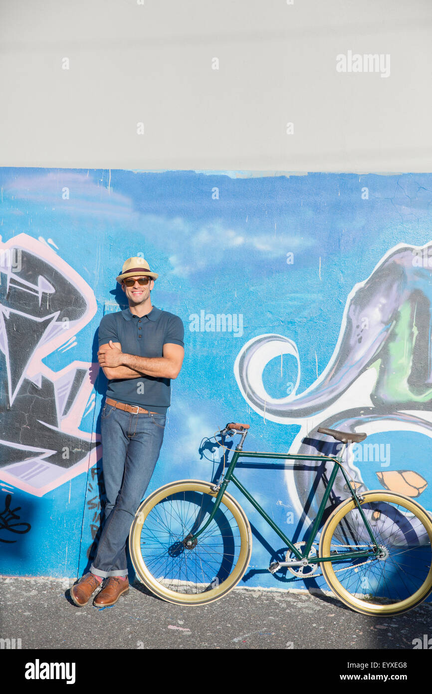 Porträt, Lächeln Mann in Hut und eine Sonnenbrille stützte sich auf urban Graffitiwand neben dem Fahrrad Stockfoto