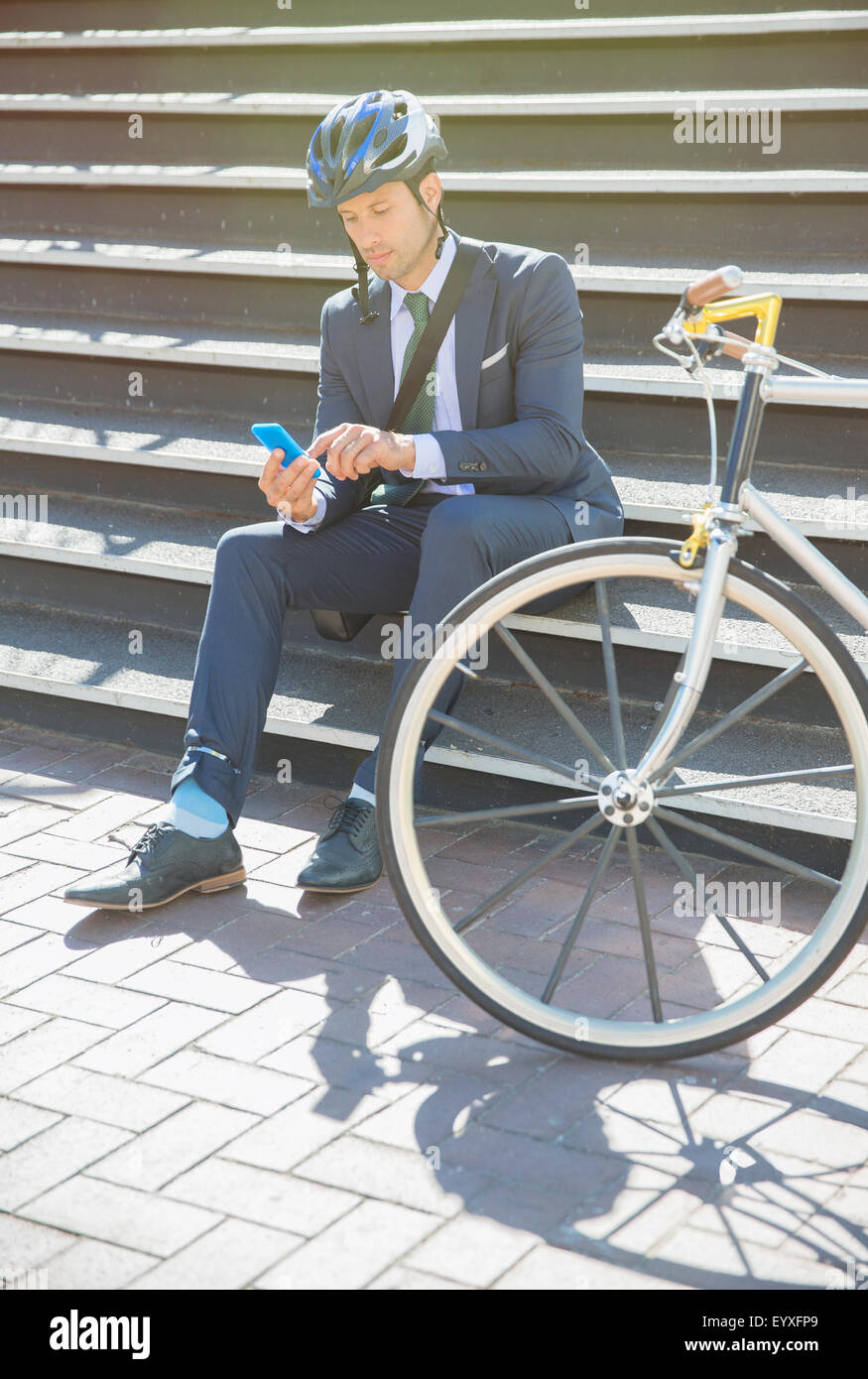 Geschäftsmann in Anzug und Helm SMS mit Handy neben Fahrrad auf sonnigen urban Treppe Stockfoto
