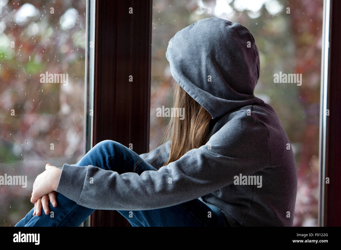 Mädchen in Kapuzenshirt, auf einer Fensterbank drinnen sitzt und nach draußen Stockfoto