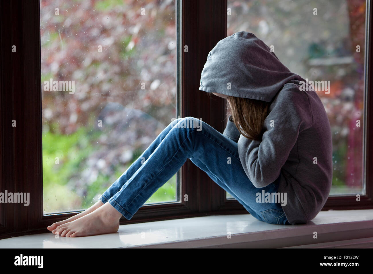 Trauriges Mädchen in Kapuzenshirt, sitzen auf einer Fensterbank mit Kopf in Händen in Verzweiflung Stockfoto