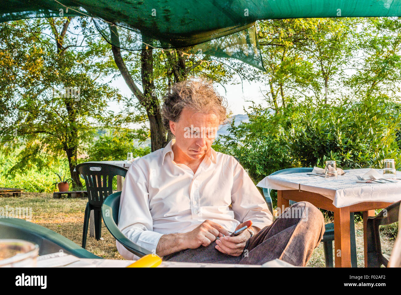 faszinierende wohlhabenden Gentleman von vierzig aus Stadt ruht auf einem Stuhl im Garten von einem roten Landhaus in Italien während mit smartphone Stockfoto
