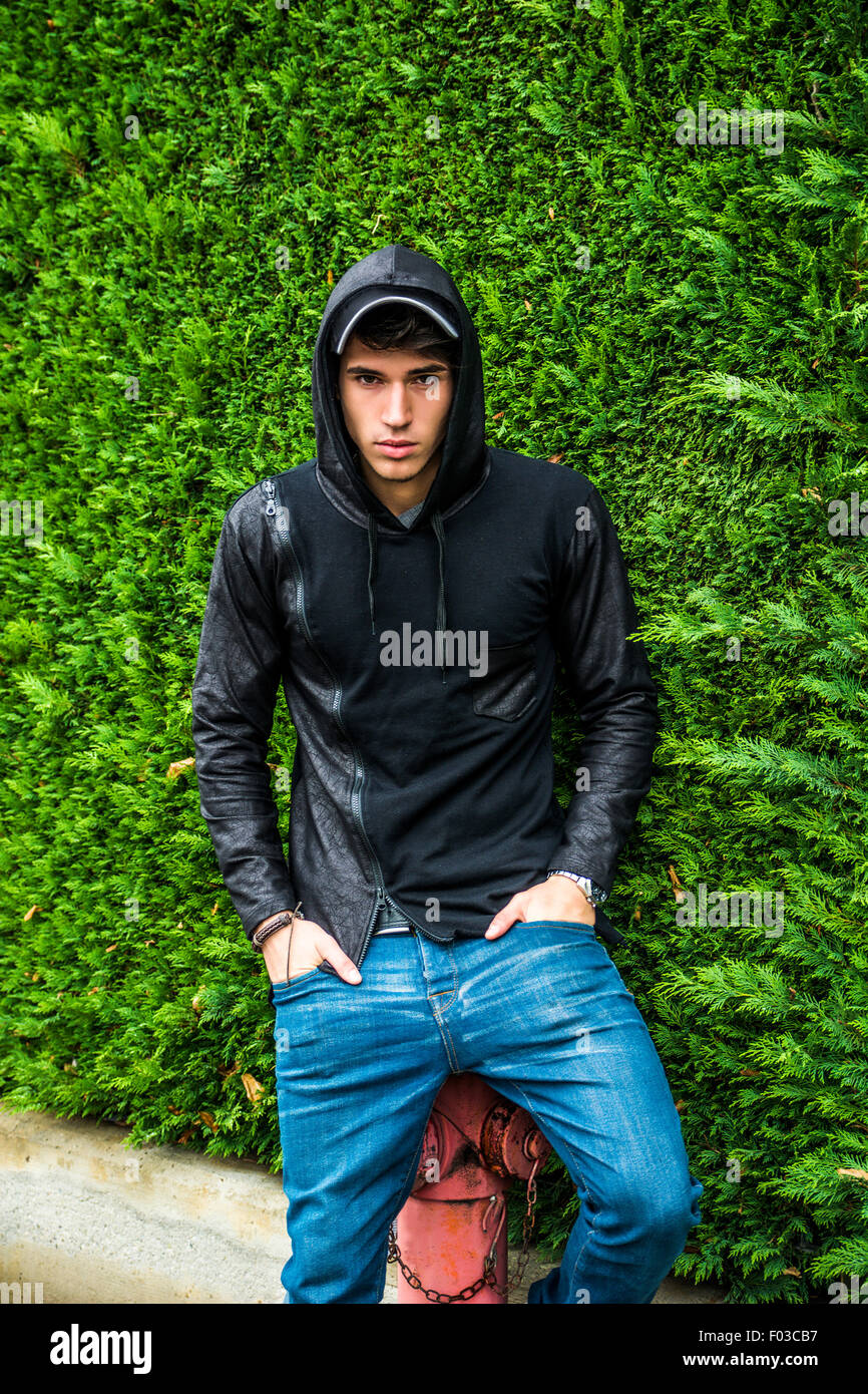 Hübscher junger Mann in schwarzer Hoodie Pullover stehen im freien gegen grüne Pflanze Hecke Blick in die Kamera Stockfoto