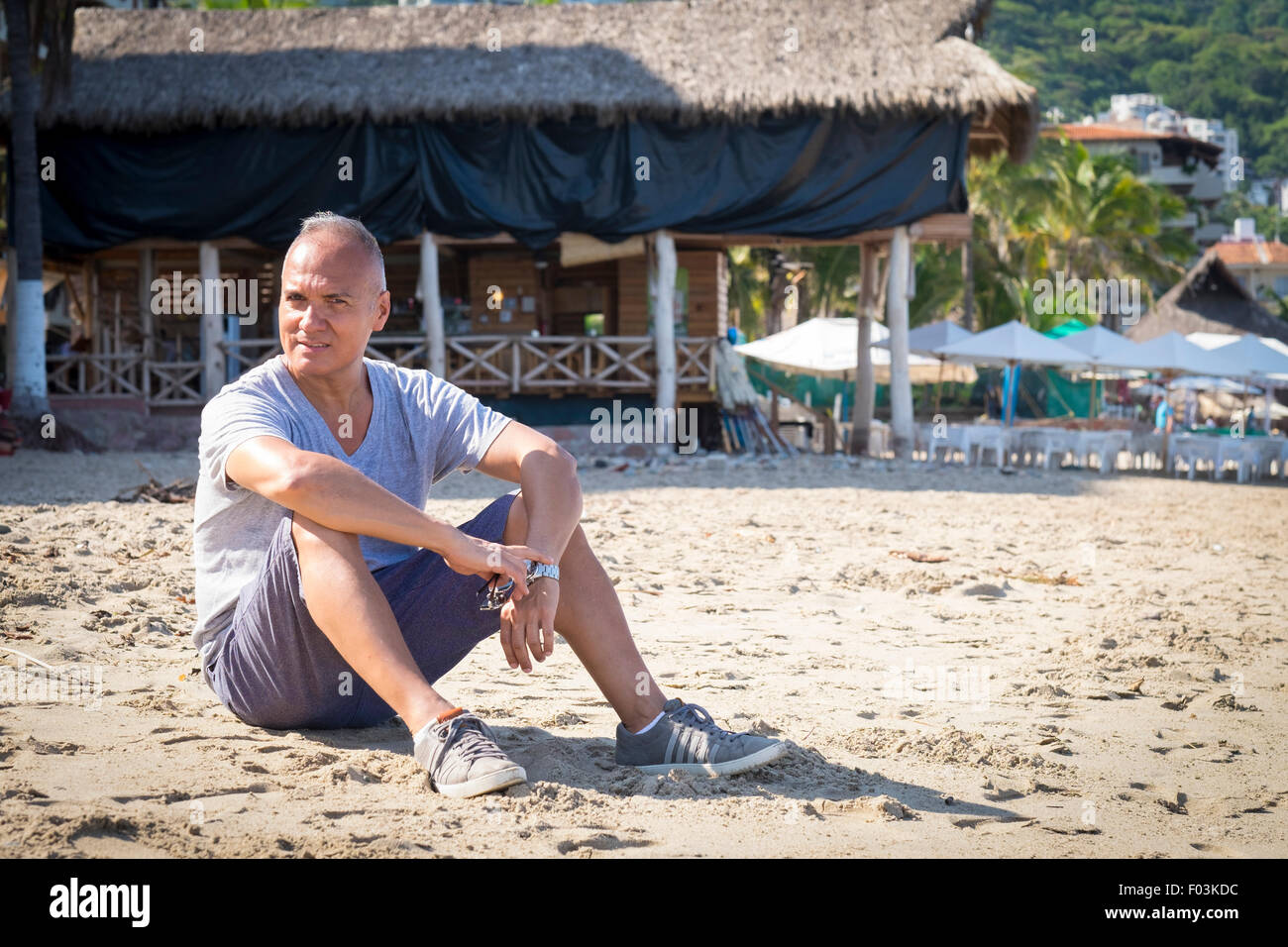 Tourismus in Puerto Vallarta, Mexiko. Mann, 55 Jahre alt, hispanische Ethnizität, im Sand sitzen. Palapa-Restaurant in der Hinterg Stockfoto