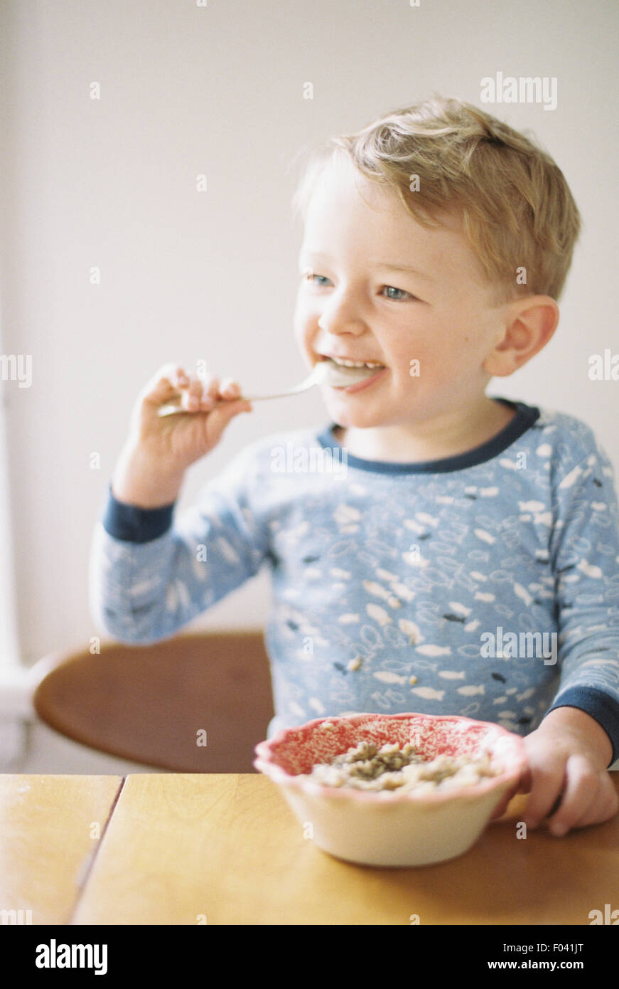 Kleiner Junge sitzt an einem Tisch beim Frühstück aus einer Schüssel. Stockfoto