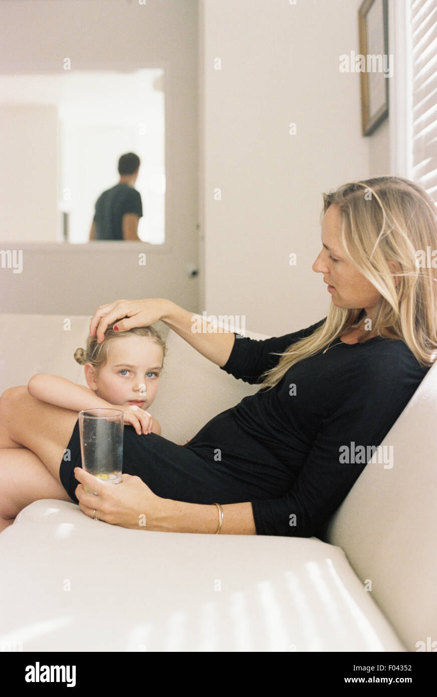 Frau mit langen blonden Haaren trug ein schwarzes Kleid, auf einem Sofa sitzt, hält ein Glas, mit ihrer Tochter zu spielen. Stockfoto