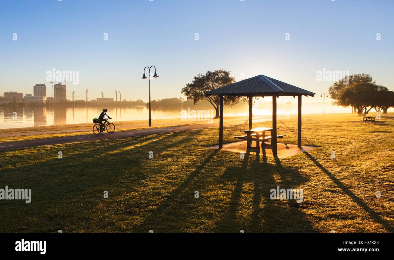 Eine Person, die mit dem Fahrrad durch Sir James Mitchell Park bei Sonnenaufgang auf einem Nebelhaften und klaren Wintertag. South Perth, Western Australia Stockfoto