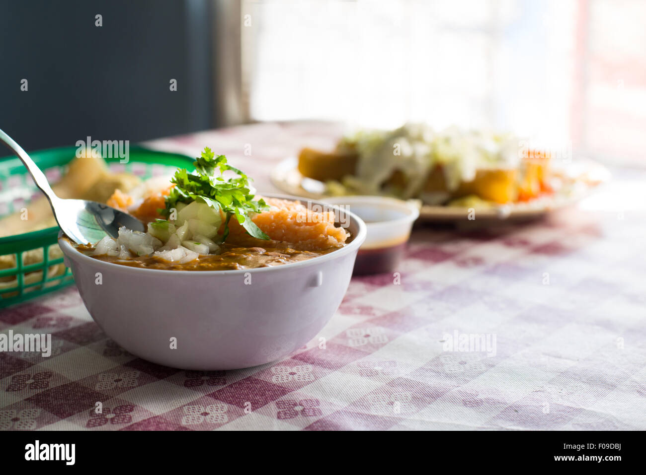 Kleine Schüssel mit Reis ein Bohnen mit Zwiebeln und Koriander garniert auf einem karierten Tischdecke mit anderen Lebensmitteln im Hintergrund Stockfoto