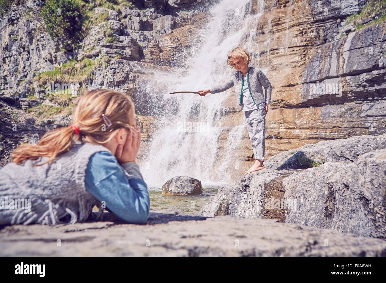 Junge Mädchen beobachten Bruder, stehend neben Wasserfall Stockfoto
