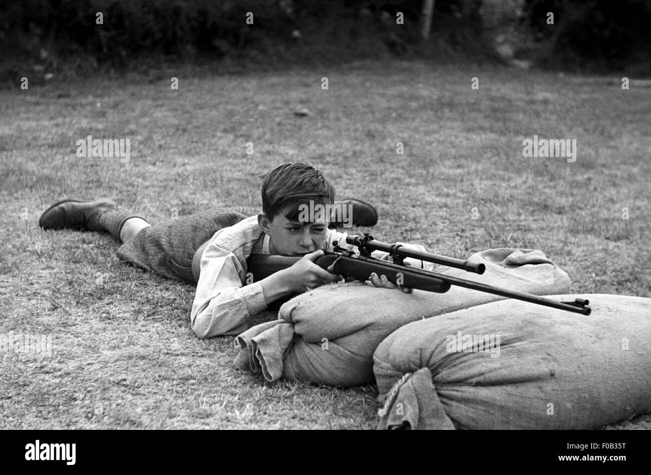 Ein kleiner Junge mit seinem Gewehr nehmen, wie er auf Sandsäcke auf dem Boden aufliegt. Stockfoto