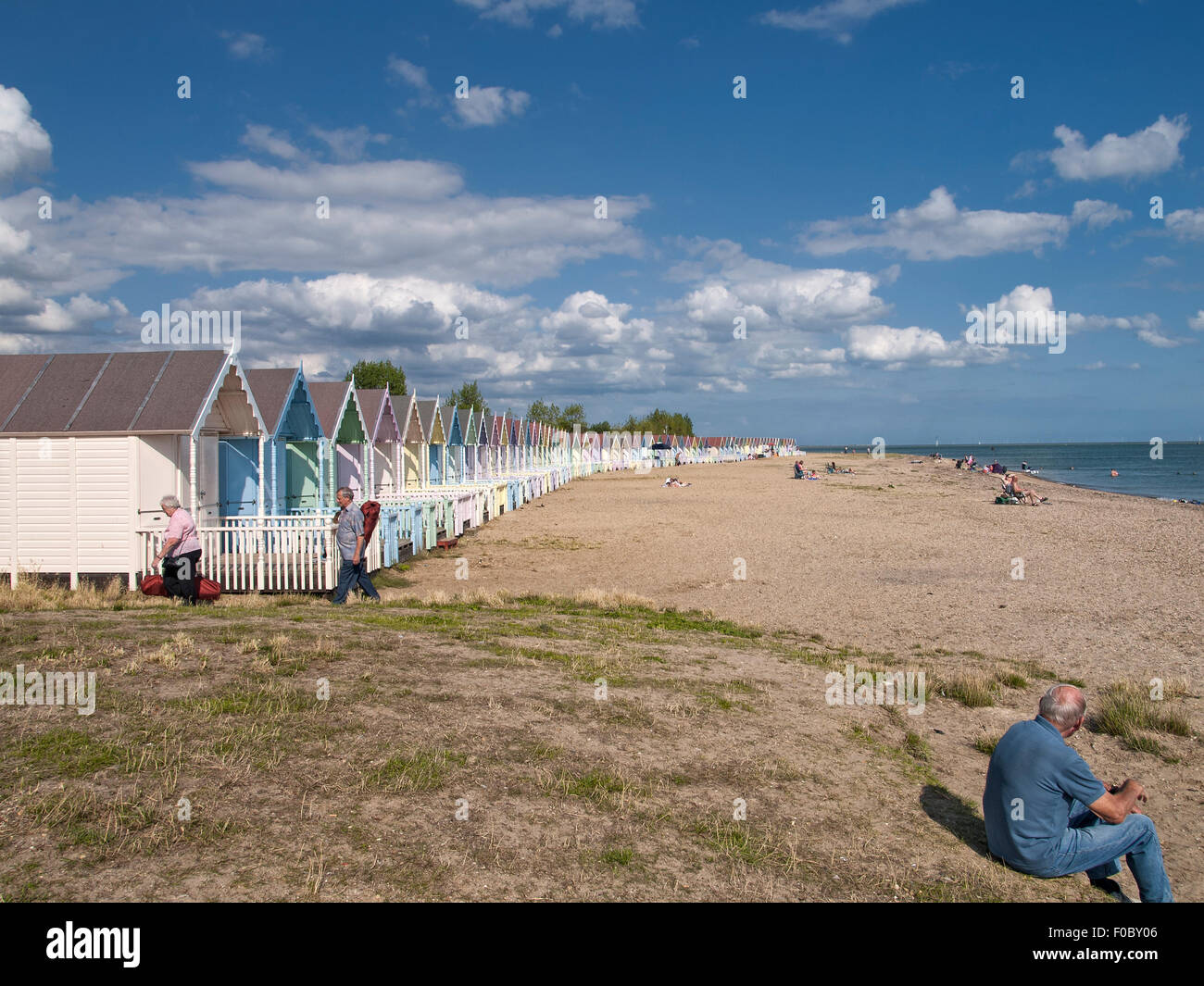 Menschen am Strand mit einer Reihe von bunten Strandhäuschen vor blauem Himmel. Mersea Island. Essex. England. VEREINIGTES KÖNIGREICH. Stockfoto