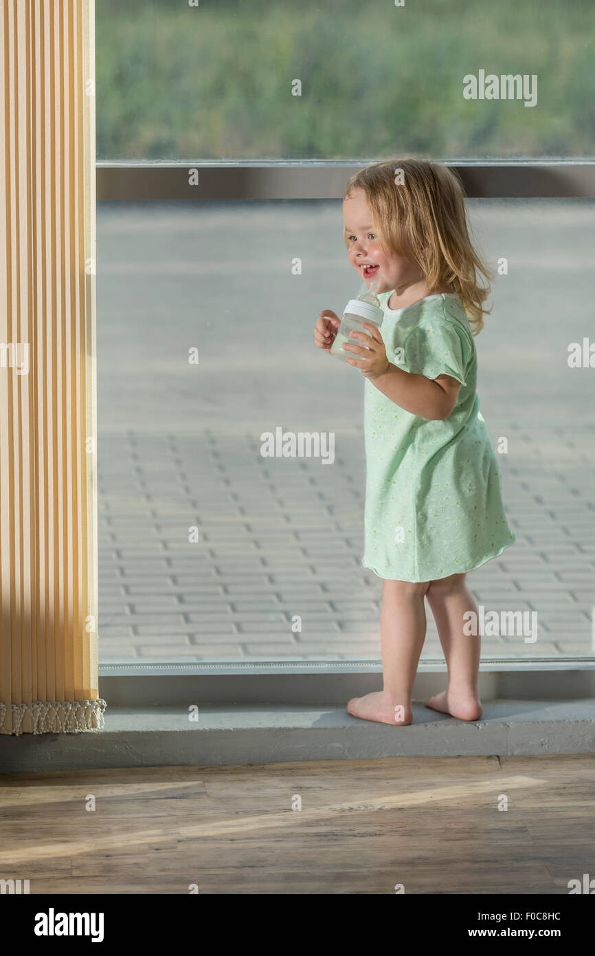 Voller Länge des Mädchens halten Babyflasche stehend durch Fenster Stockfoto