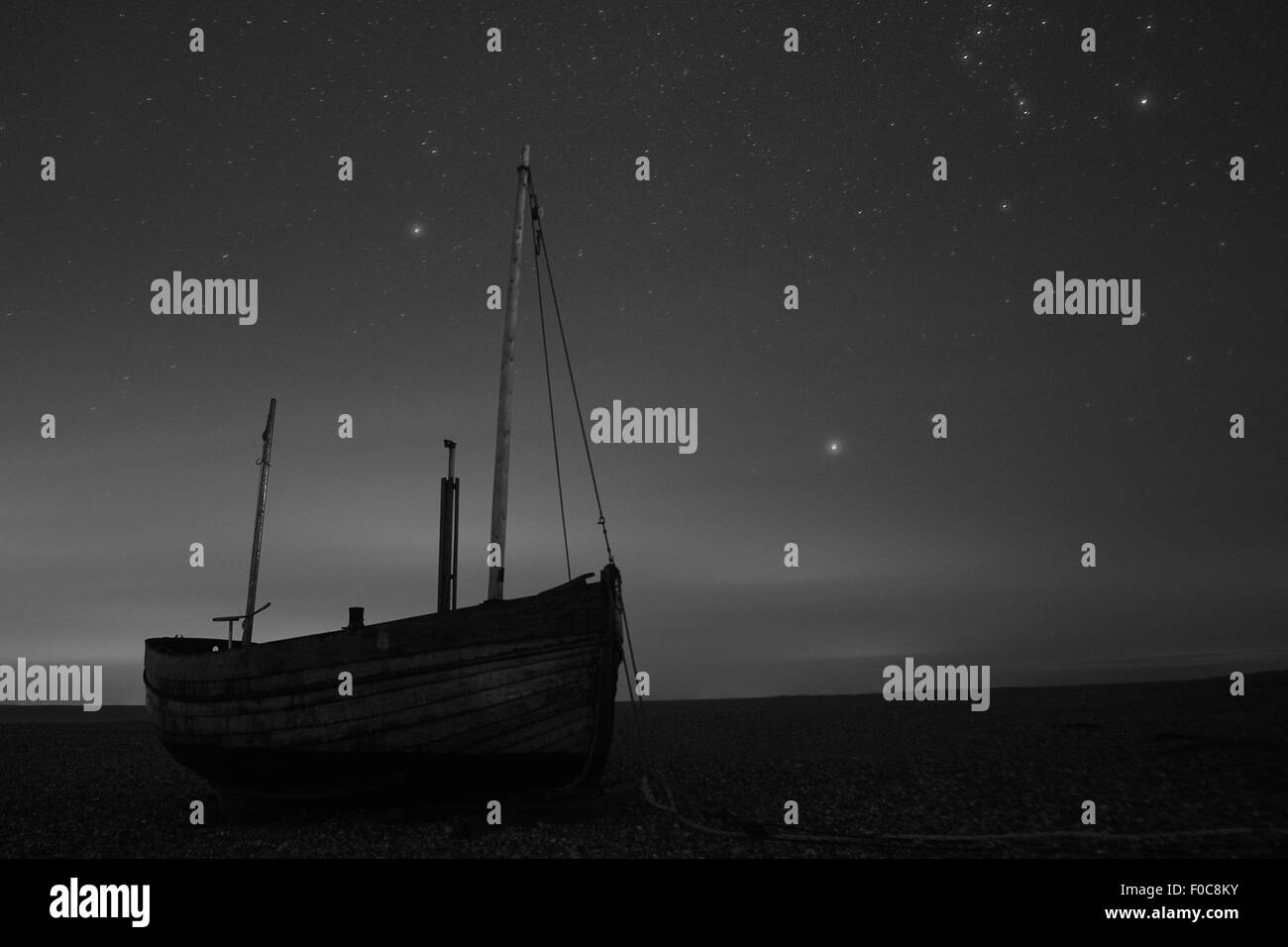 Ein hölzernes Boot sieht man am Strand in der Nacht unter dem Sternenzelt, in Dungeness, Kent, England, in schwarz und weiß. Stockfoto