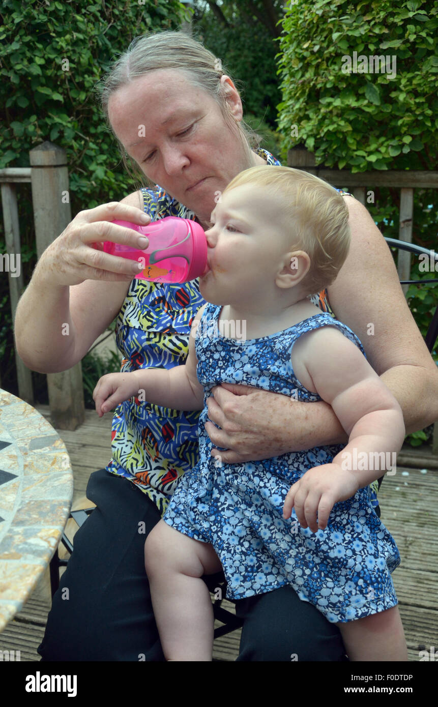 Eine Großmutter (oder ältere Mutter) hält ihr zehn Monate altes Enkelkind (oder Baby) Wer ist Wasser aus einem Becher trinken. Stockfoto
