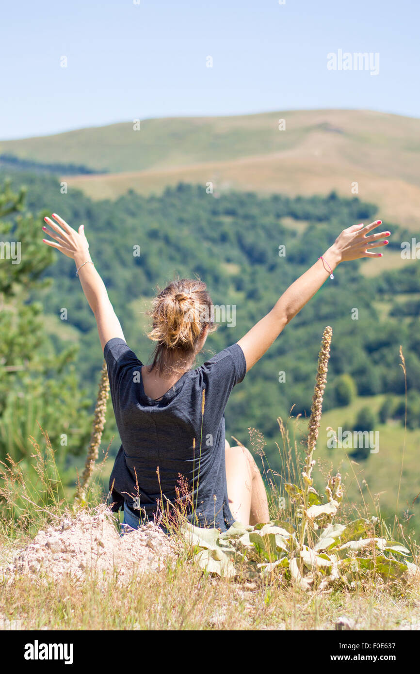 Junge Frau mit Rucksack sitzt am Rande der Klippen und Blick auf Berge und Himmel mit erhobenen Händen. Wandern Reise Freude Stockfoto