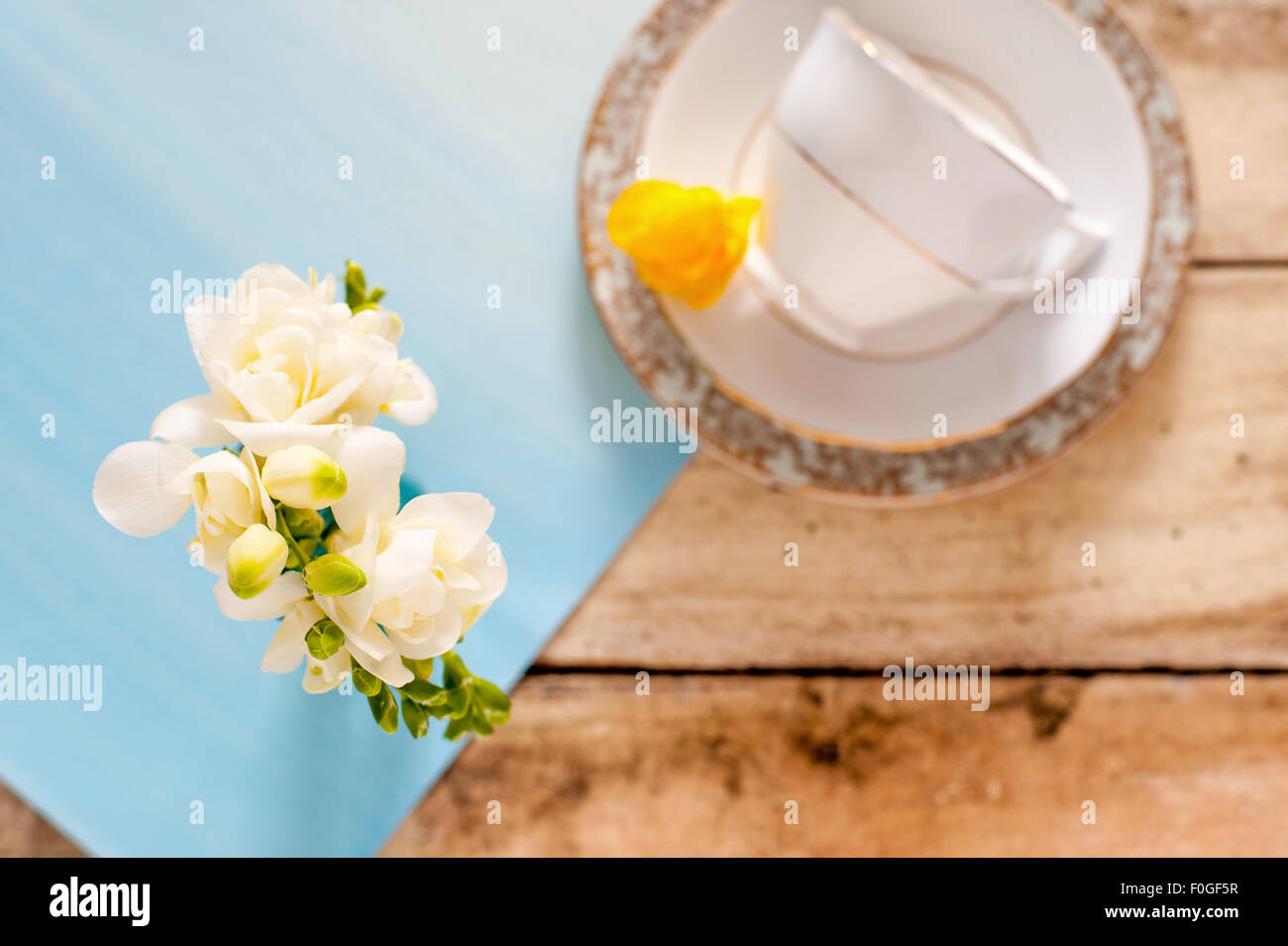Nahaufnahme des weißen Freesien auf blauen Tischset mit unscharfen Vintage Teetasse und Untertasse im Hintergrund, auf rustikalen Holztisch Stockfoto