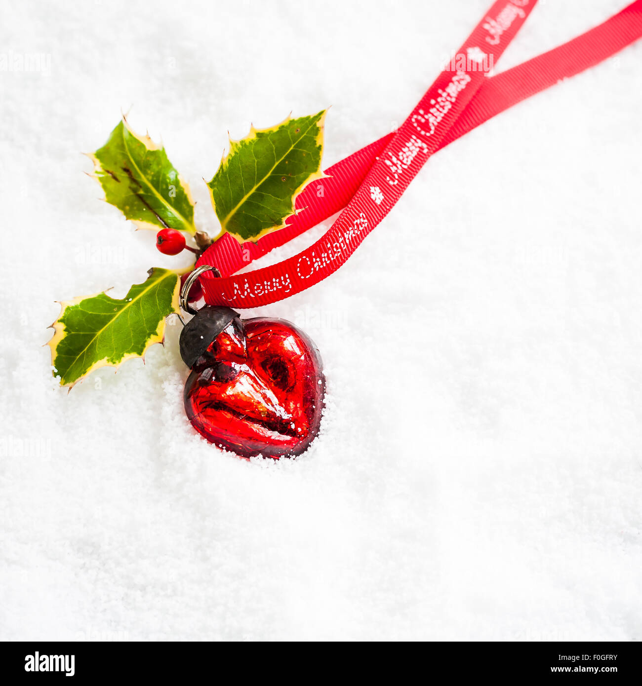 herzförmige Rote Christbaumkugel mit Holly und Frohe Weihnachten Ribbon auf Schnee Stockfoto