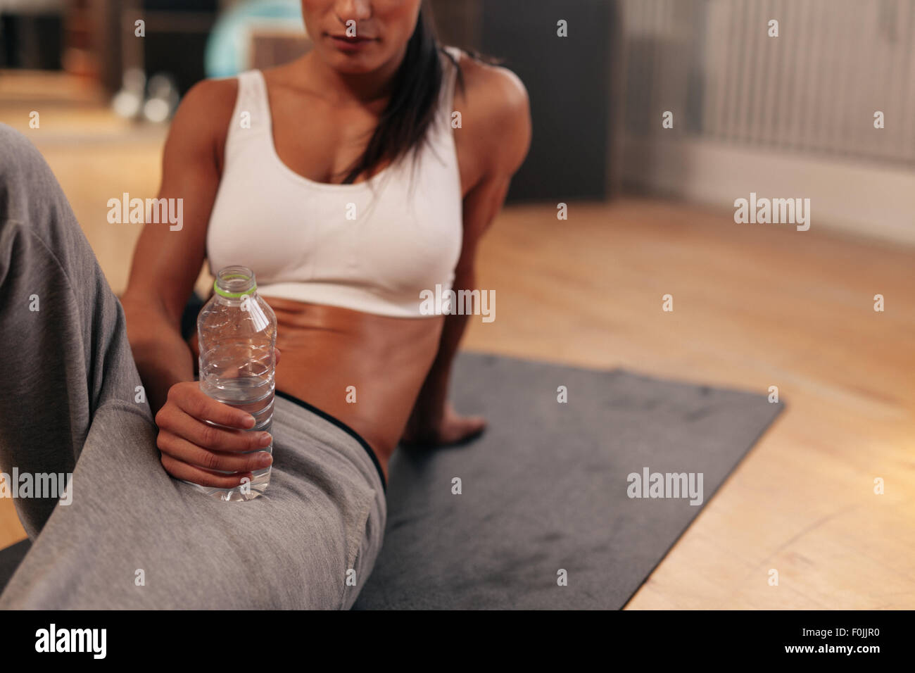 Schuss entspannte junge Frau mit Flasche Wasser abgeschnitten. Fitness-Frau im Sport tragen auf Gymnastikmatte in Turnhalle sitzen. Stockfoto