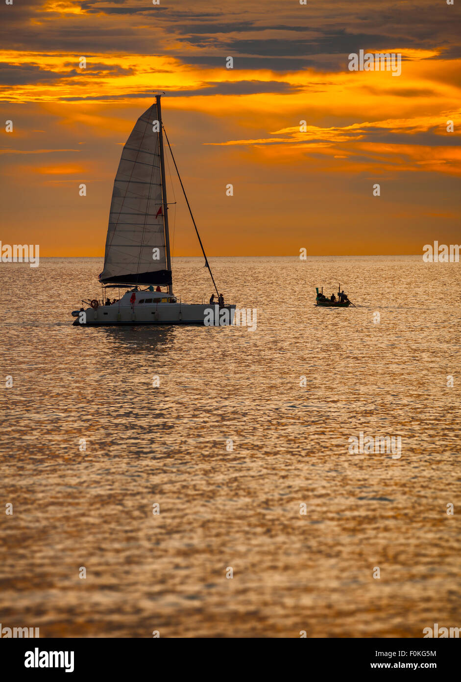 Himmel lodernden mit dem letzten orange Licht des Sonnenuntergangs über ein Katamaran Segelboot, mit seiner Segel entfaltet, auf einem tropischen Meer. Stockfoto