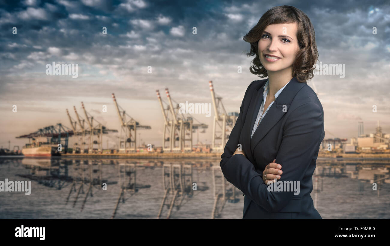 Attraktive Geschäftsfrau mit lockigen braunen Haaren trägt eine modische Jacke stand vor einem Hafen oder Business Port Ansicht smil Stockfoto