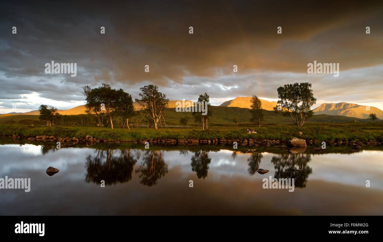 Goldenes Licht trifft moor die Hügeln oberhalb von Rannoch von der untergehenden Sonne mit einer Reflexion der dunklen Wolken und ein Regenbogen in Loch Ba Stockfoto