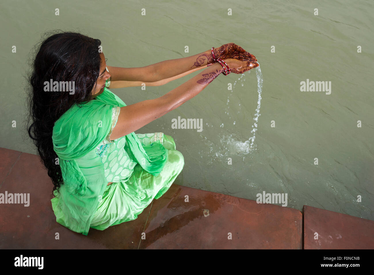 Eine junge Frau mit langen schwarzen Haaren, Henna bemalte Hände und ein grünes Kleid ist an den Ghats von dem heiligen Fluss Ganges beten. Stockfoto